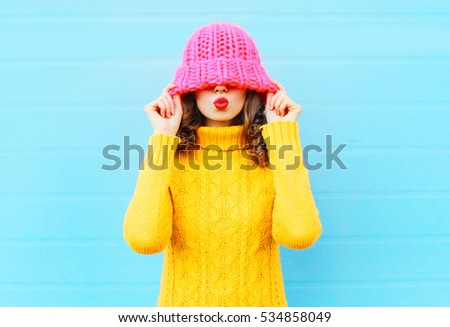 Fashion happy woman blowing red lips makes air kiss wearing a knitted hat, yellow sweater over blue background