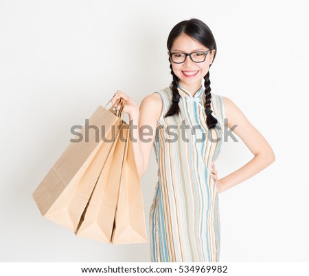 Portrait of young Asian woman in traditional qipao dress shopping, hand holding paper bag, celebrating Chinese Lunar New Year or spring festival, standing on plain background.