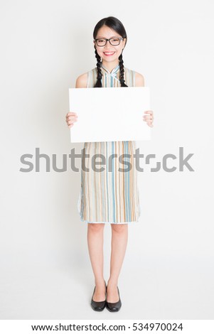Portrait of young Asian woman in traditional qipao dress hand holding a white blank paper card, celebrating Chinese Lunar New Year or spring festival, full body standing on plain background.