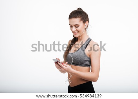Portrait of a smiling fitness woman texting on mobile phone and wearing earphones isolated on a white background