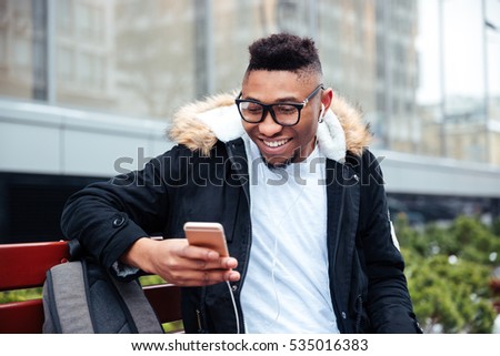 Picture of african cheerful man holding his cellphone in hands and chatting while listen to music outdoors.