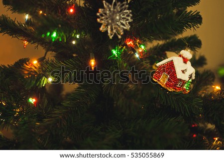 Toy house on a christmas tree. Decorating home before holidays. Warm and cozy photo with green pine tree, garland and christmas lights