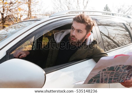 Picture of losted in forest bearded shocked driver sitting in car while holding map. Looking aside.