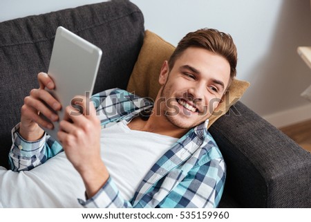 Picture of a smiling bristle man dressed in shirt in a cage print lies on sofa in home and using tablet computer. Looking aside.