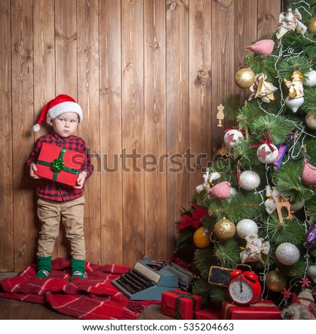 Smiling funny child boy in Santa hat opening Xmas gifts. New Year and Christmas miracle. Christmas family traditions. Happy Holidays home with family. Wood Fence Background with copy space. Flat lay