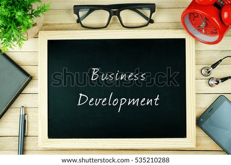 Top view of pen, diary, small green tree, spectacle, alarm clock, earphone, smartphone and black chalkboard written with BUSINESS DEVELOPMENT inscription. Business conceptual.