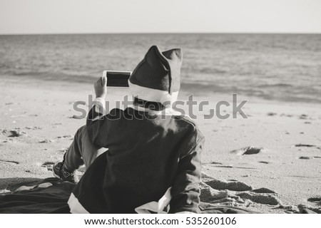 Back Side View of Joyful Child Santa Claus Using Smart Phone on Sunny Sky Ocean Beach Outdoors Background. Mock up screen, playing game on winter tropic holiday vacation. Black and white picture