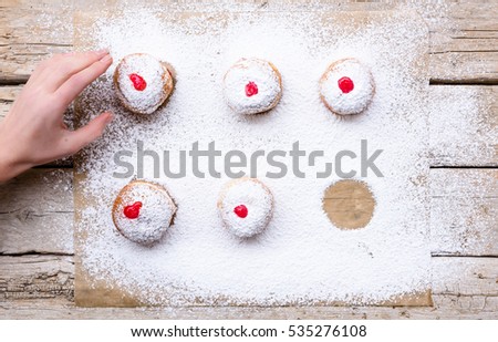 Fresh donuts (sufgania) with red jelly jam on a sugar powder and wooden background. Hand of woman taking donut from bakery paper. Hanukkah holiday celebration and traditional jewish sweet