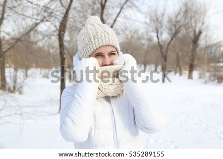 The girl's portrait in the winter in a white down-padded coat, a knitted cap and mittens against the background of snow of trees in the city park