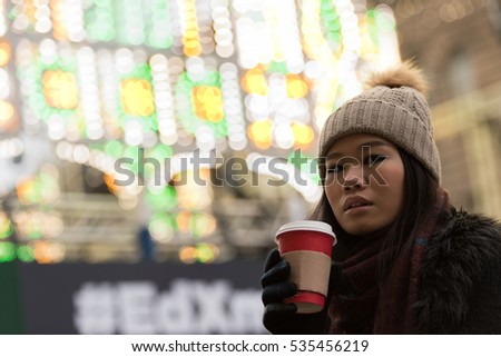 An Asian girl wearing a woolly hat, scarf and gloves stands in front of a light display holding a warm cup of coffee as she looks away in George Street, Edinburgh City Center, UK.