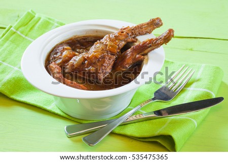 Braised rabbit with onions in beer, in a white ceramic bowl with checkered napkin on a green wooden table, horizontal