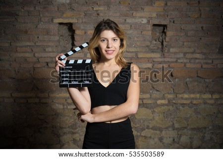 Young blonde woman actress in dark basement, smiling face expression with movie clapper board, looking at camera