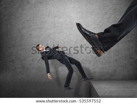A giant foot in a black shoe kicking a little businessmen off the edge, and he is falling down, on the concrete gray background. Major defeat. Bankruptcy and insolvency. Failing business. Royalty-Free Stock Photo #535597552