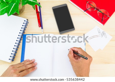 Hand writing on notebook on wooden desk with office supplies, smartphone, glasses and green leaves pot, business concept
