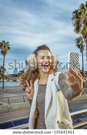 in Barcelona for a perfect winter. happy young woman in earmuffs in Barcelona, Spain  taking selfie with smartphone