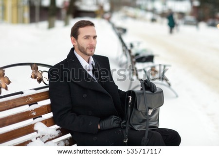 Male businessman sitting on a bench, holding a business briefcase with documents, in the winter on the street