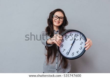 Smiling cute Asian woman in glasses and gray shirt holding clock in hands and looking at camera. Focus on clock. Isolated gray background