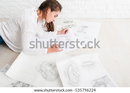 Successful woman artist drawing in workshop. Talented painter painting sketches of human portraits with pencil, lot of pictures around. Art, craft, inspiration, intensive work concept