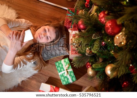 Top view of a beautiful woman holding her phone in her hands, surrounded by christmas presents. Girl lying on floor with fir tree branch. Young woman checking out her text messages on her mobile phone