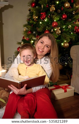 A little fair haired boy and his mom reading a book together laughing for a picture sitting under a christmas tree. Mom with child reading book and relaxing by the fire place some cold evening.