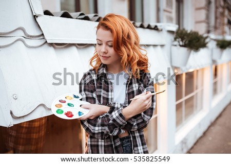 Picture of incredible young lady painter with red hair walking on the street. Look at palette while holding paintbrush outdoors.