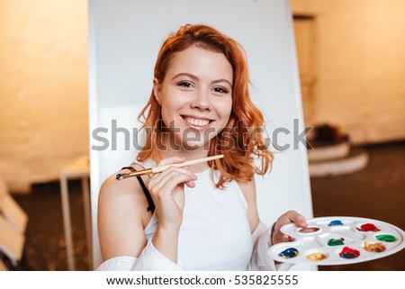 Picture of happy redhead young woman painter standing over blank canvas in artist workshop. Look at camera while holding palette and paintbrush