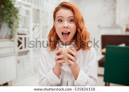 Picture of young redhead surprised happy woman drinking coffee in cafe. Looking at the camera.