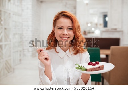 Picture of redhead young happy woman eating cake in cafe. Looking at the camera.