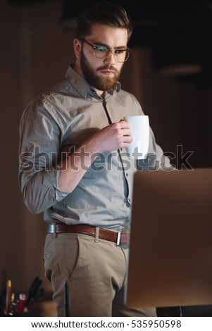 Picture of concentrated designer dressed in shirt and wearing eyeglasses working late at night while drinking tea.