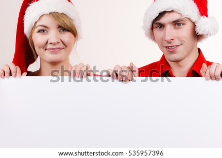 Couple holding banner sign with copy space for text, peeking over edge of blank empty billboard. Happy glad girl and boy in santa claus hats. Christmas advertisement copyspace.