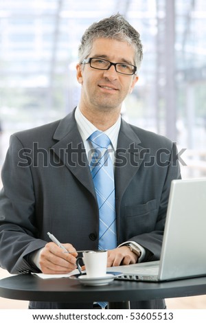Businessman writing notes on paper standing at coffee table in lobby of corporate building, in front of windows.