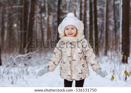 little three year old smiling girl with blond hair and gray eyes, a beige knitted hat and scarf, knitted mittens, grey and beige jacket, is in a white snow-covered Park or forest in the winter.