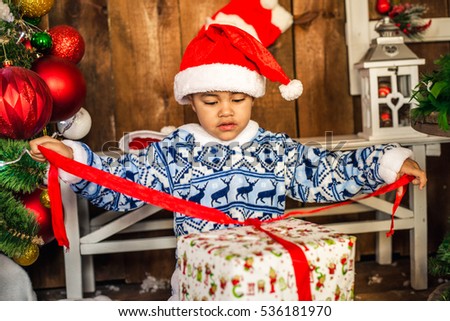 Little boy opening a Christmas present. African-American boy in the hat of Santa Claus opens Christmas gift