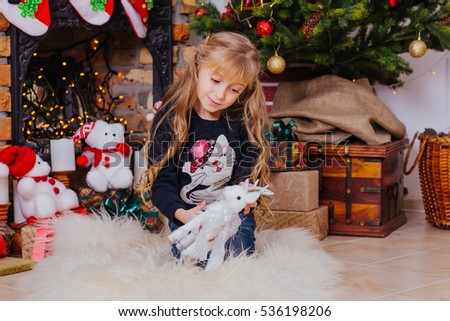 portrait of a little delicate girl who is sitting under the Christmas tree in a stylish sweater and jeans, holding a toy and looks at toy