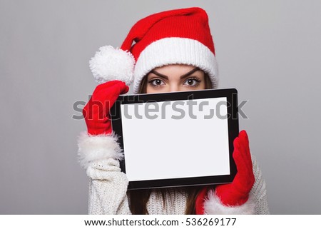 Beautiful young Caucasian woman in sweater, gloves and Santa knit hat looking at camera, holding digital tablet with blank screen. Copy space, no retouch, studio lighting.