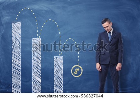 Businessman standing upset and a column diagram with a dollar sign falling down painted on the blackboard behind him. Currency falling. Crisis effect. Money loss. Royalty-Free Stock Photo #536327044