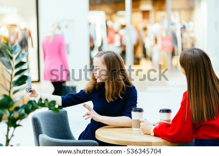 Two young attractive girls sitting on the table in cafe and taking selfie in shopping center. 