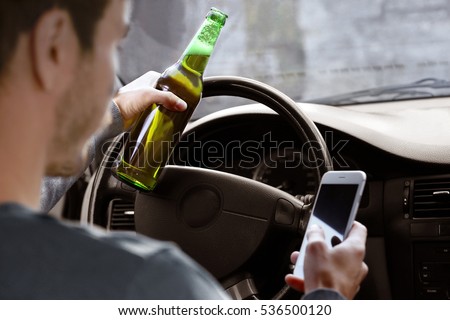 Man holding mobile phone and bottle of beer while driving car, closeup. Don't drink and drive concept Royalty-Free Stock Photo #536500120