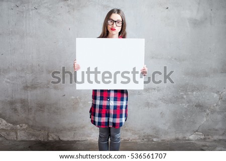 Young woman dressed casually holding white board or empty placard with copyspace standing on the gray wall background.