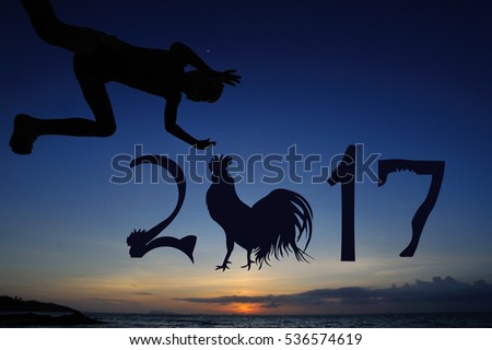 Silhouettes on the beach with a background of sunset sky: little boy jumps from dad's hands and flutters over the water. There are numbers 2017 written in the air - year of the rooster is coming up