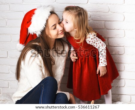 young cute little blonde girl in christmas red dress kissing her smiling mother in xmas hat and long brunette hair on white brick wall background