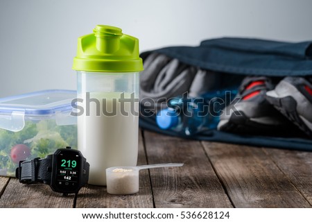 Must have of every sportsman in the foreground of the camera. Special sports watch with important functions, shaker with protein, container with salad made of greenery and bag with clothes and shoes. Royalty-Free Stock Photo #536628124