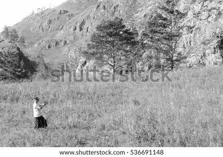 Black and white photograph of a male tourist in a wide pants and a sailor taking pictures of nature on camera at a hill with trees near the mountain Altai. Soft focus.