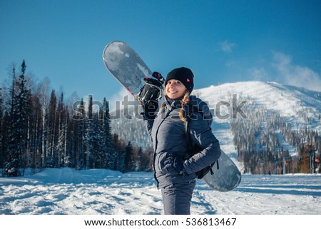 Beautiful young blond woman in pigtails  in grey winter costume smiles, stands and holds in hand snowboard on a background of ski resort and descent from the snow mountain at sunny day