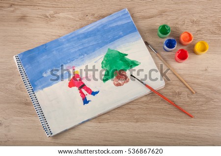 art workplace. children christmas picture on paper of forest with Santa, pine tree and colored paints, brushes on wooden table. top view