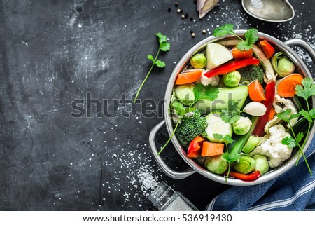 Various fresh vegetables in a pot - colorful fresh clear spring soup (vegetarian bouillon or stock). Kitchen scenery from above (top view). Black chalkboard background - layout with free text space.