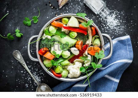 Various fresh vegetables in a pot - colorful fresh clear spring soup (vegetarian bouillon or stock). Cooking - kitchen scenery from above (top view). Black chalkboard background.
