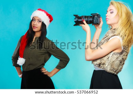 Photographer and model. Caucasian blonde girl shooting images, taking photos with camera., photographing african woman in santa helper hat.