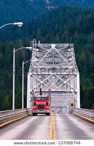 Red semi truck rides on the road with a yellow stripe dividing across the silver metal bridge on the background mountains covered with dense forest