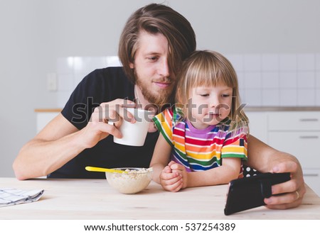 young father drinking morning coffee and watching mobile phone together with kid on kitchen 